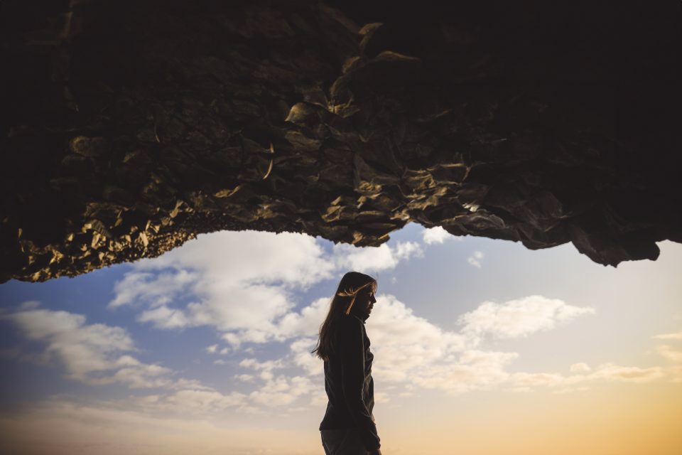 A solitary silhouette of a person wanders beneath a rugged rocky overhang, framed by a moody, cloud-laden sky.