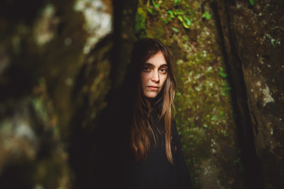 A person with long hair stands in a dimly lit, rocky area, surrounded by moss-covered rocks, as if nature's own auto draft had sketched this serene landscape.