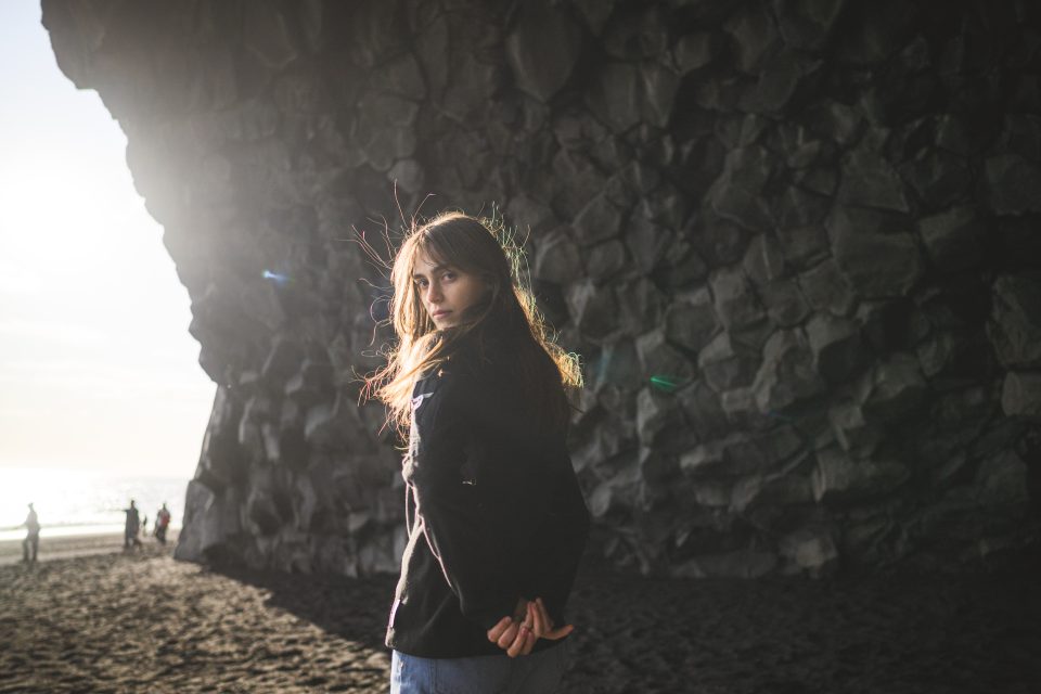 A person with long hair stands on a rocky beach, facing the camera. Sunlight streams in from the left, like an artist's auto draft, highlighting the textured cliff in the background.