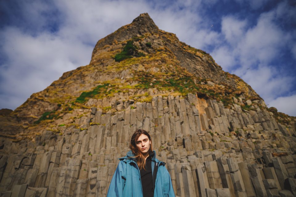 A person in a blue jacket stands in front of a rocky, columnar basalt formation under a sky with clouds that seem like an ethereal auto draft of nature’s own script.