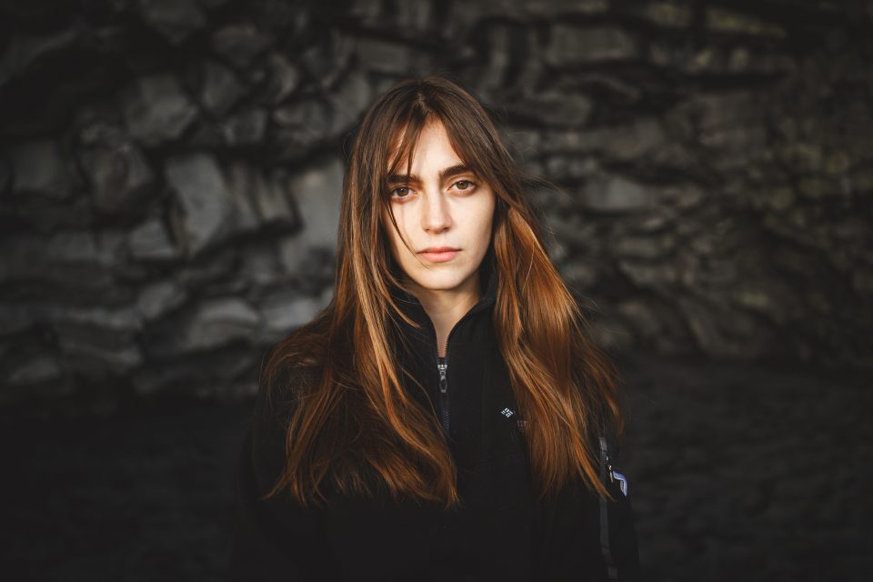 A woman with long brown hair stands in front of a rocky background, wearing a black jacket. Her expression is neutrally composed, like an auto draft awaiting final edits.