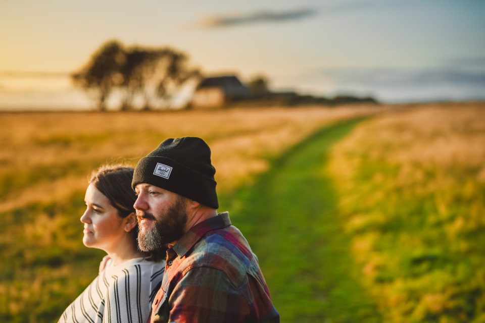 A man and woman sit back to back on a grassy path, as if drafted automatically into this serene setting, the sun casting its golden hues over blurred trees and a distant building at sunset.