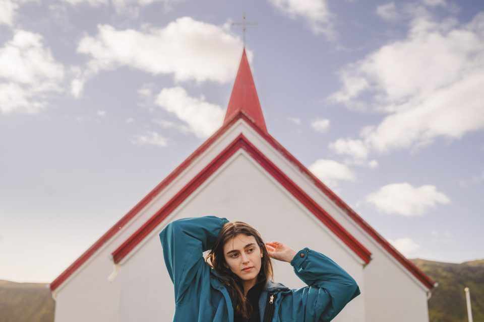 A person in a teal jacket stands in front of a red and white church with a pointed steeple under a partly cloudy sky, almost as though painted by an artist capturing the scene in an auto draft.