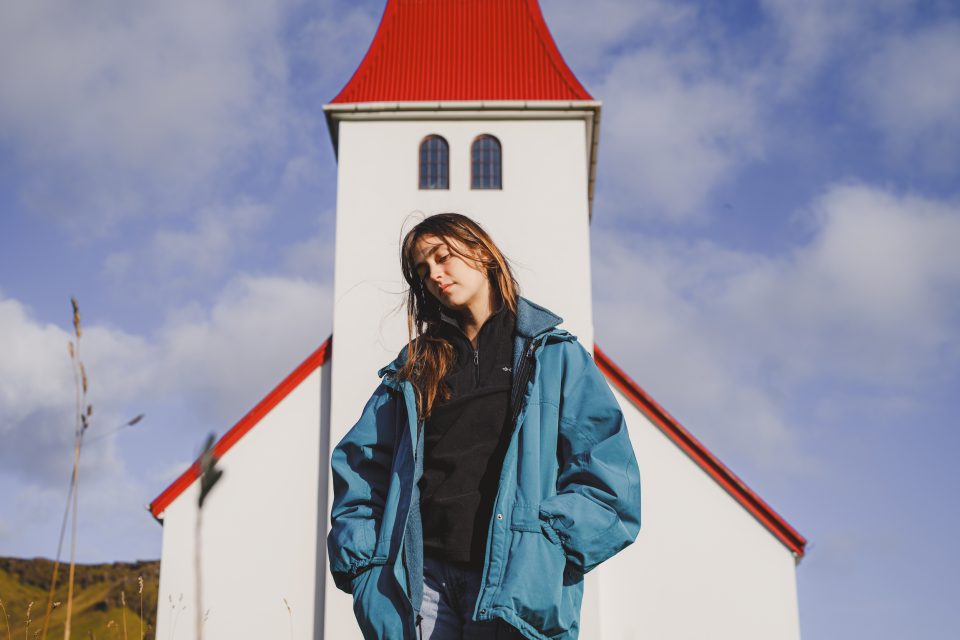 A woman in a blue jacket stands gracefully in front of a picturesque white church with a striking red roof, all set against a dramatic cloudy sky.
