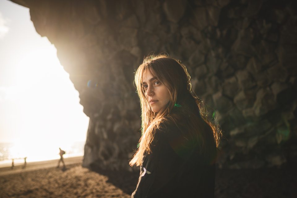 A person with long hair stands on a beach at sunset, their silhouette perfectly framed by the rock formation, as if nature itself penned this breathtaking auto draft.