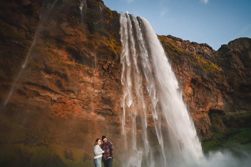 A couple stands near a majestic waterfall cascading down a rocky cliff, basking in the refreshing mist under a clear blue sky.