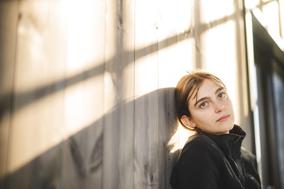 A person with short hair and a black jacket leans against a wooden wall, where sunlight casts intricate shadows, creating an atmosphere filled with mystery and intrigue.