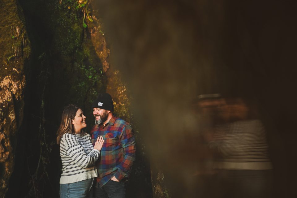A man and woman stand close together outdoors, both smiling as if capturing an auto draft of their perfect moment. The background is blurred with greenery and shadows.