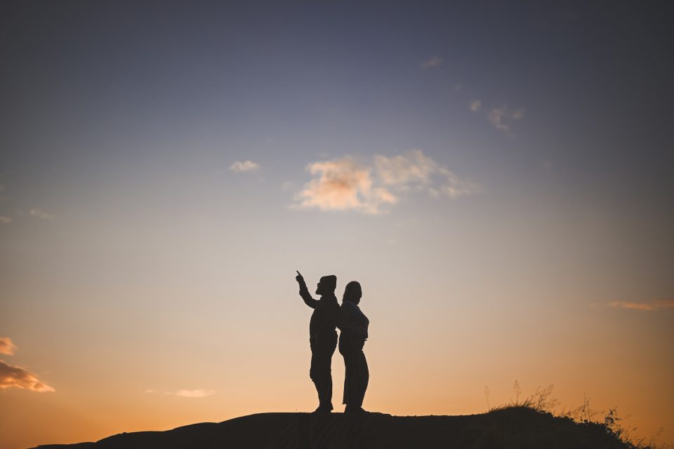 Silhouettes of two people stand on a hill, one pointing into the distance. The sky, like an artist's auto draft, shifts from orange to purple, suggesting sunset.