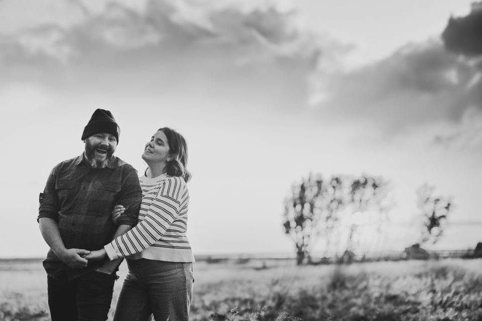A couple stands in a scenic field, holding hands and smiling, framed by lush trees and a cloudy sky.
