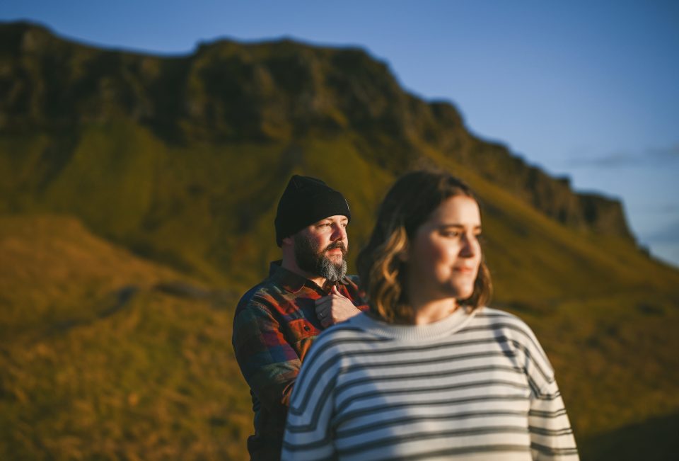 Two people stand outdoors in front of a grassy hill and rocky formation. The person in the foreground, clad in a striped shirt, seems like an artist sketching an auto draft from nature, while the person behind wears a beanie and plaid shirt, observing the serene landscape.