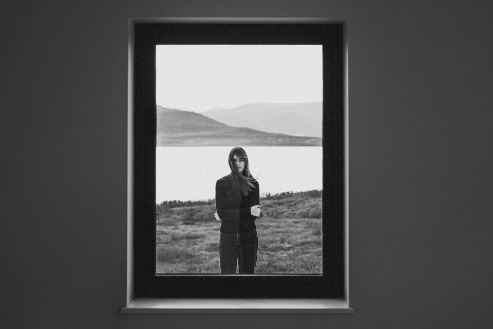 A woman stands outside, framed by a window, with a lake and hills in the background. The image captures the timeless elegance of her silhouette in black and white, as if drafted like an auto sketch against nature's serene backdrop.