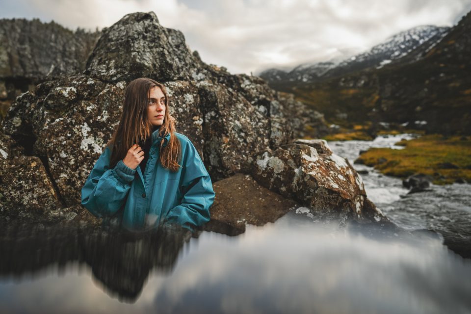 A woman in a blue jacket sits on rugged rocky terrain near a flowing mountain stream, reflecting under the moody, cloudy sky.