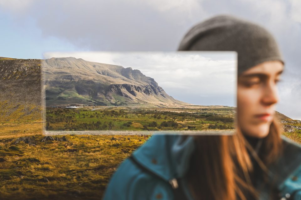 A young woman in a beanie and blue jacket stands confidently in front of a scenic, auto draft-like mountainous landscape, partially blurred in the foreground.