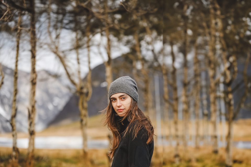 A woman, looking thoughtfully at an auto draft of her latest novel, wears a gray beanie as she stands in front of a row of trees, with majestic mountains forming the backdrop.