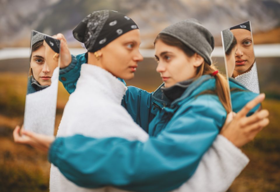 Two people in outdoor attire hold mirrors reflecting each other's faces, like an auto draft of nature's artwork, against a blurred natural background.