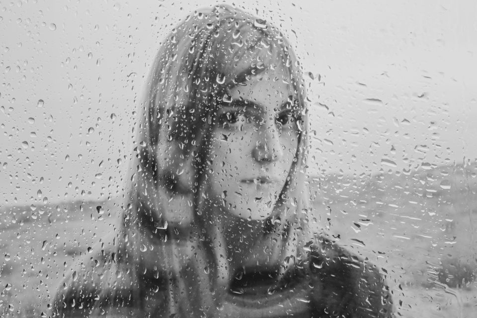Through the rain-streaked window, a person with long hair gazes into the distance, their silhouette a study in contrasts. The black and white image evokes a sense of longing, reminiscent of an auto draft left unfinished yet full of potential.