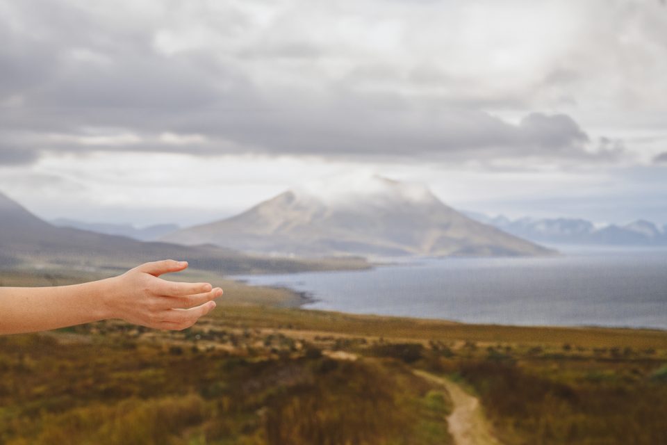 A hand reaches out towards a cloudy mountain landscape, with a winding path resembling an auto draft leading to a distant body of water.
