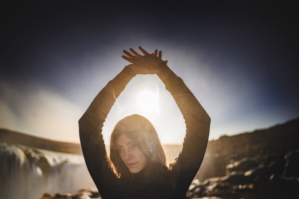 A person stands, arms raised, against the backdrop of a waterfall and bright sun, their silhouette perfectly framing this scene that feels like an auto draft of nature's grand design.