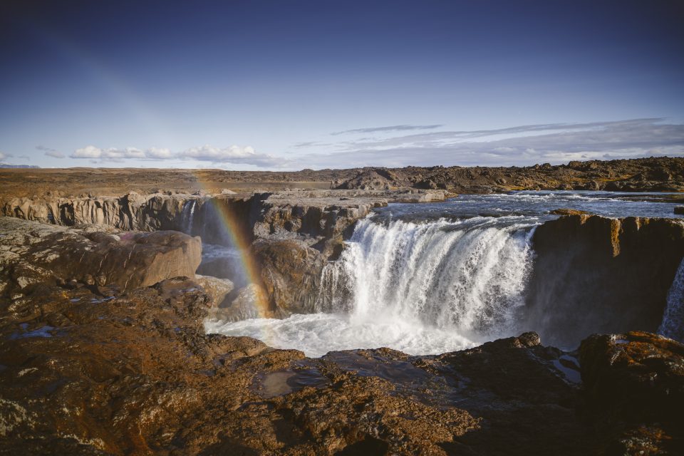 A wide waterfall with a rainbow stretching across it, captured through the refined lens of a Nikon Z8. Rocky terrain surrounds the scene under a clear blue sky, making it perfect for any photography review.