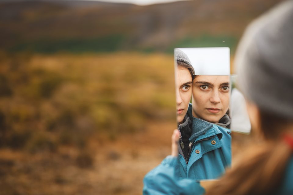 A person holds fragmented mirrors, each shard catching glimpses of their face against a blurred outdoor background, reminiscent of an auto draft capturing fleeting moments.