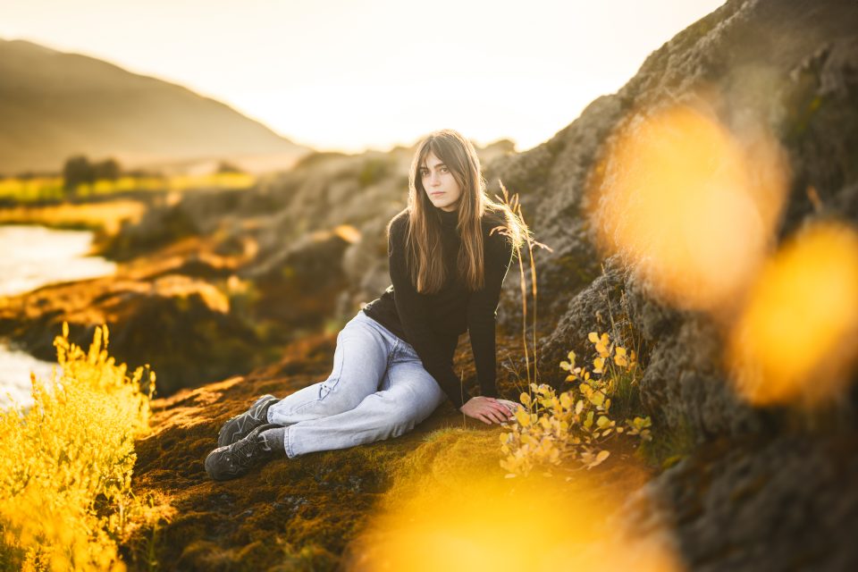 A woman in a black sweater and jeans sits on a rocky terrain beside a shimmering body of water, surrounded by yellow foliage, with mountains in the background, as if capturing nature's auto draft for her soul.