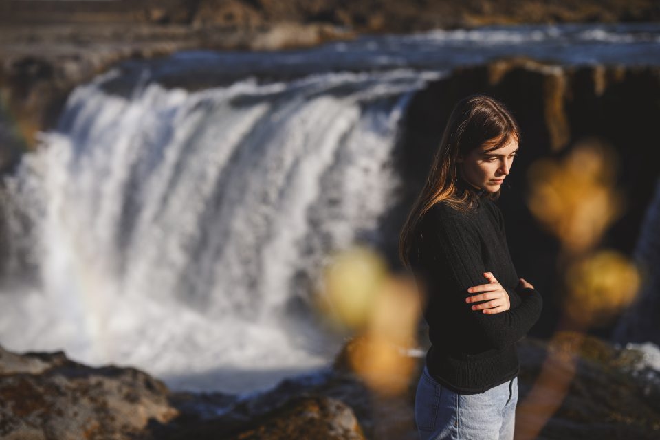 A person stands with crossed arms, as if auto-drafting their next adventure, in front of a large waterfall with sunlight casting soft shadows.