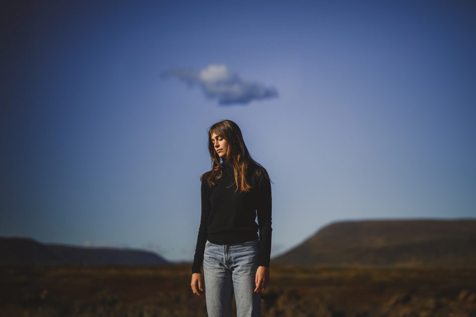 A woman in a black sweater and jeans stands outdoors under a blue sky with a single cloud, resembling an auto draft of nature's artistry. Hills are visible in the background.