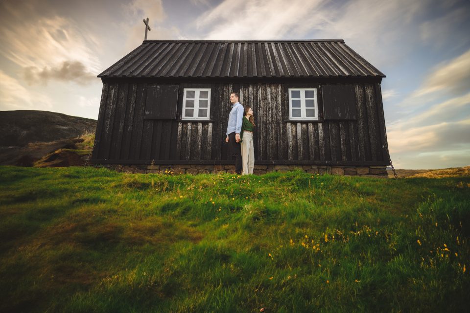 Two people stand in front of a rustic black wooden cabin with a slanted roof and cross on top, amidst a grassy landscape under a partly cloudy sky. It feels like the setting for an auto draft of adventure, where stories begin to write themselves.