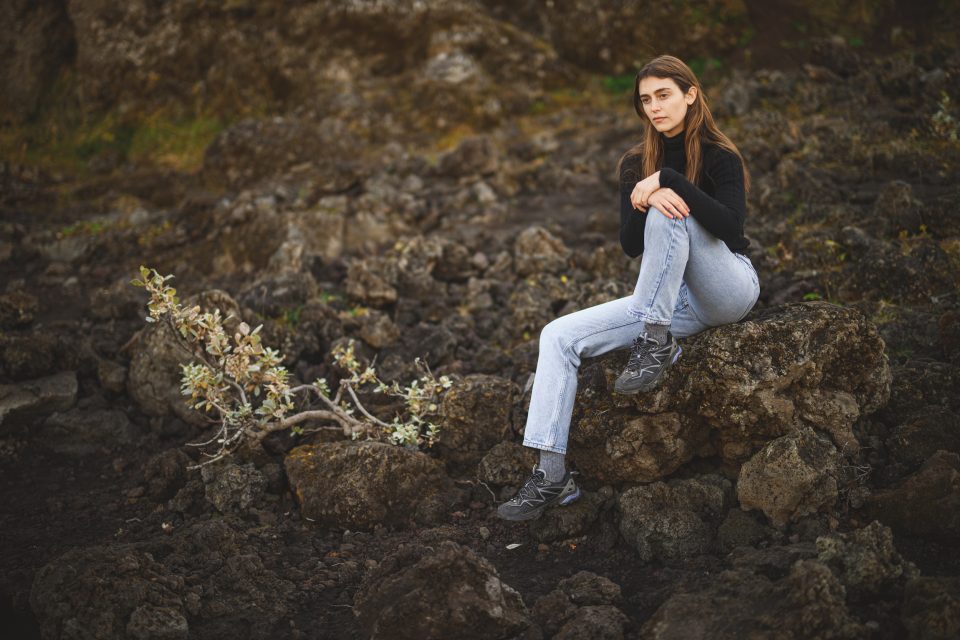 A person sits on a rocky terrain, wearing a stylish black top and jeans, resembling an Auto Draft model in nature, with a small plant nearby.