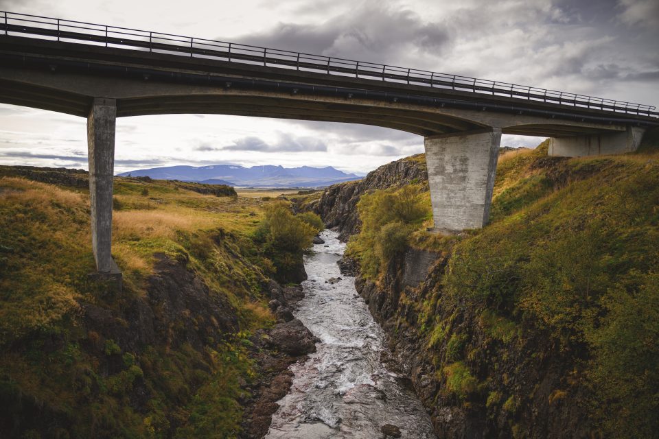 A concrete bridge, resembling an auto draft of architectural precision, spans a narrow, rocky river gorge surrounded by grassy hills and distant mountains under a cloudy sky.