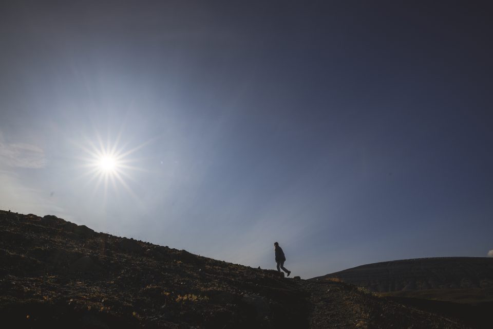 Under the bright sun in a clear sky, a person is walking uphill with determination, framed by the majestic mountain backdrop—it’s an experience one wouldn't want to auto draft into memory.