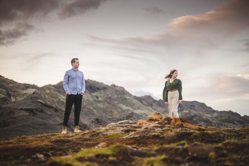 A man and a woman stand on a rocky hillside, framed by mountains in the background under a cloudy sky, as if pausing to draft their next adventure.