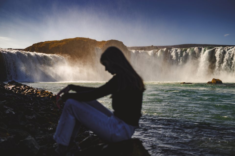 A silhouetted person sits by a rocky shore, contemplating the auto draft of nature's masterpiece—a large waterfall cascading in the background under a clear blue sky.