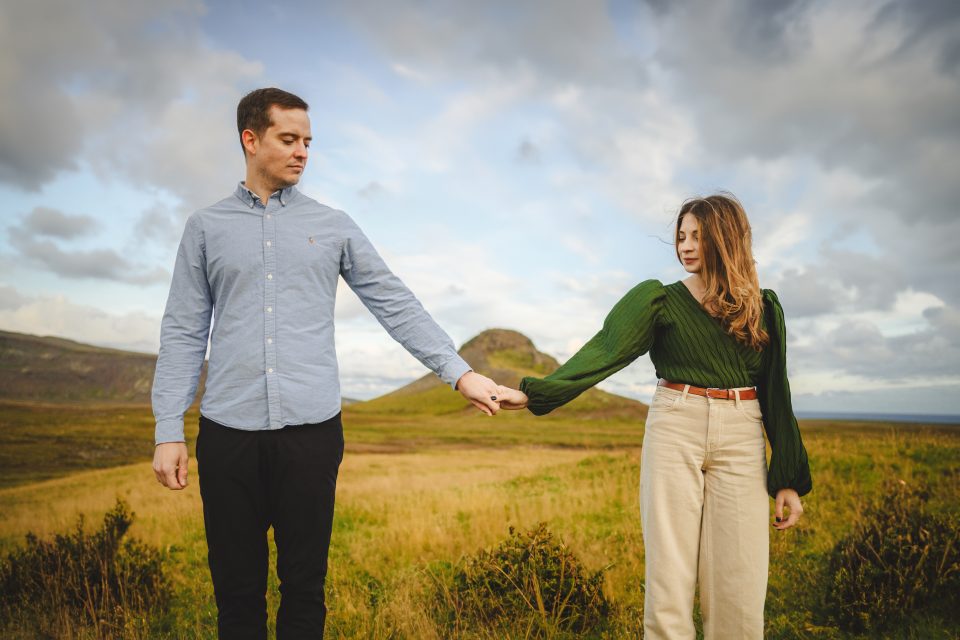 A man and woman stand hand in hand in a grassy field, beneath a cloudy sky with rolling hills in the backdrop, capturing an enchanting moment that defies the mundane.