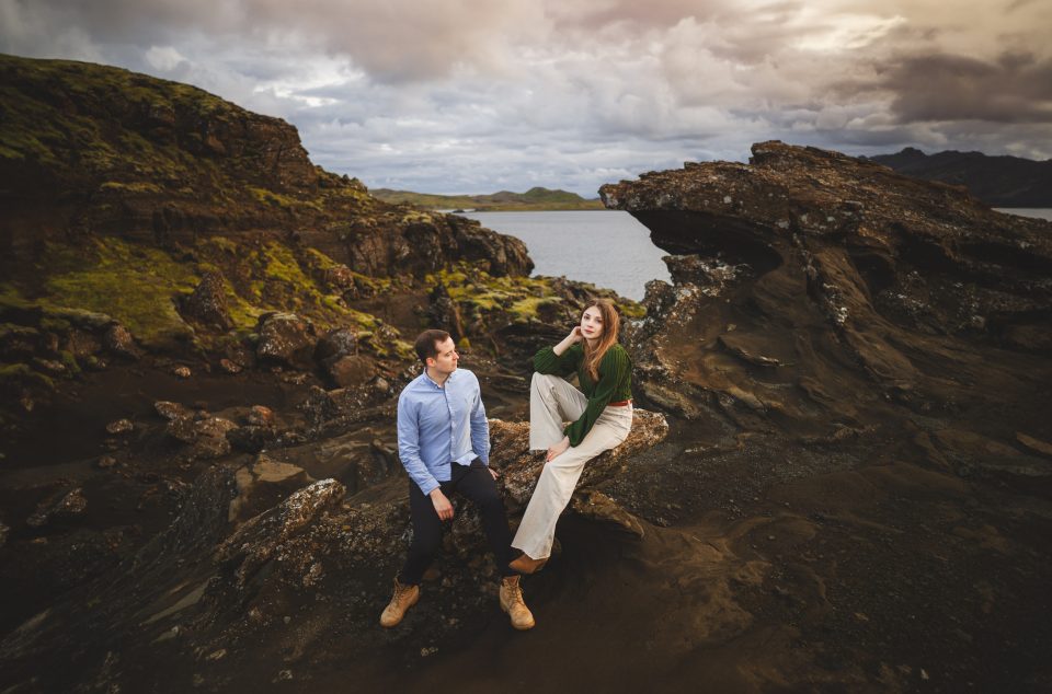 Two people sit on a rocky outcrop near a lake, sketching with an automated drafting tool, surrounded by rugged terrain and cloudy skies.
