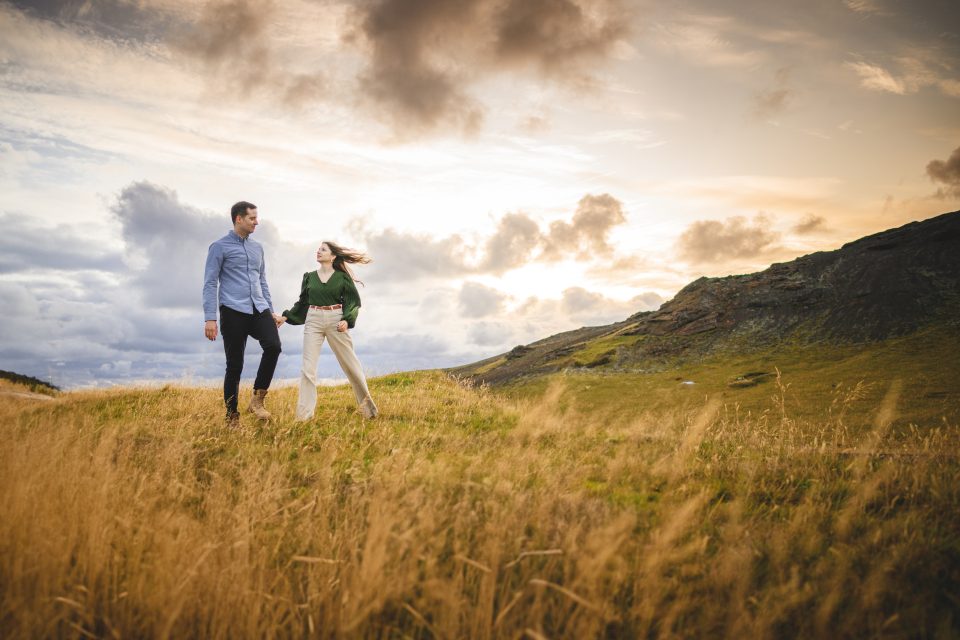 A couple walks hand in hand through a grassy meadow at sunset, with golden hills and a dramatic, cloud-filled sky serving as nature's backdrop. Their journey feels like a well-composed auto draft, capturing the perfect moment amidst the serene beauty of an evening's embrace.