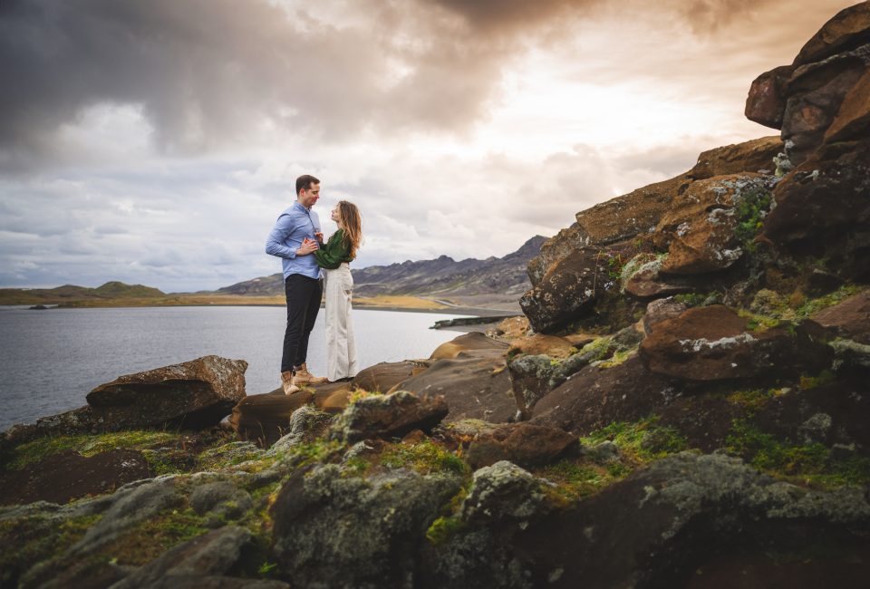 A couple stands on rocky terrain near a vast body of water, their gaze as intense as the dramatic, cloudy sky above—a scene worthy of an artist's auto draft, capturing nature's raw emotion.