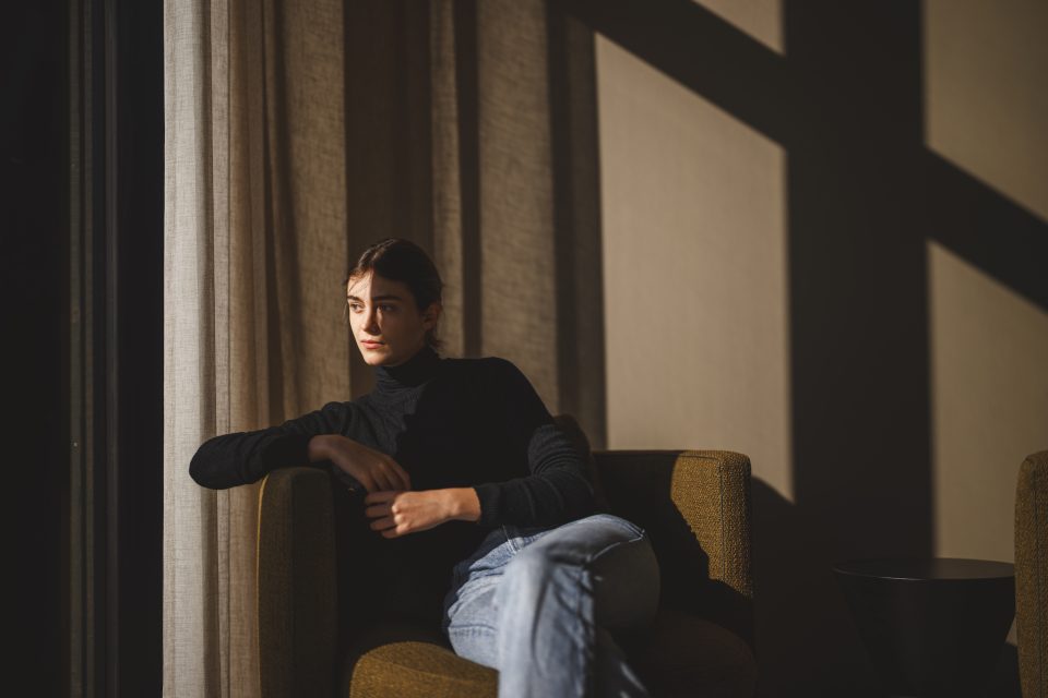 A person in a dark sweater sits by a window, bathed in sunlight, shadows cast on the wall like an artist’s auto-draft of light and darkness.