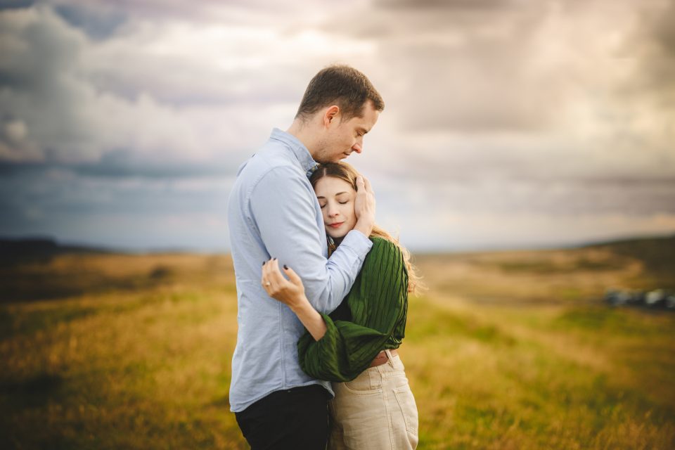 A couple embraces in a field under a cloudy sky, the dry grass whispering softly in the gentle breeze.