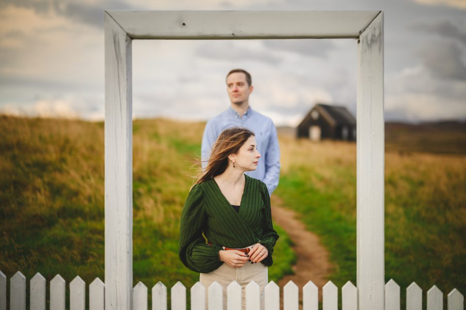 In the foreground, a woman stands framed by a white fence, with a man positioned behind her. The scene captures the timeless feel of an auto draft on canvas, set against a backdrop of a grassy field and small house.