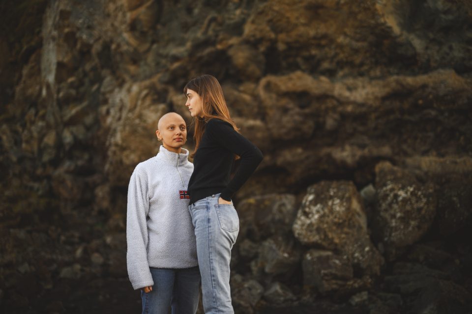 Two people stand in front of a rocky background like an auto draft sketch coming to life. One person in a white sweater faces the camera while the other in black looks to the side, capturing a perfect moment of contrast and intrigue.