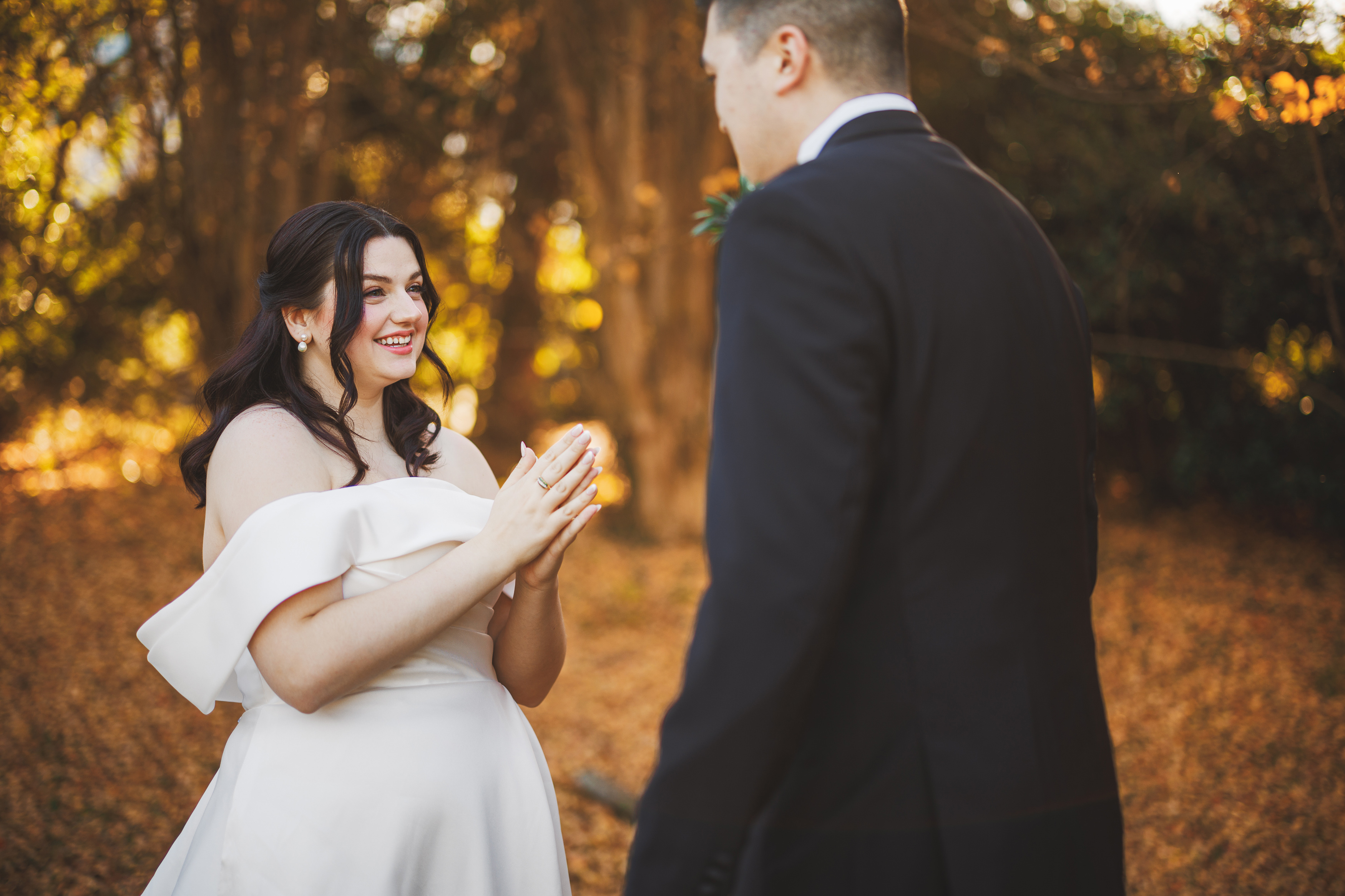 A woman in a white dress smiles with hands clasped, facing a person in a suit. They stand outdoors with trees and autumn leaves in the background.
