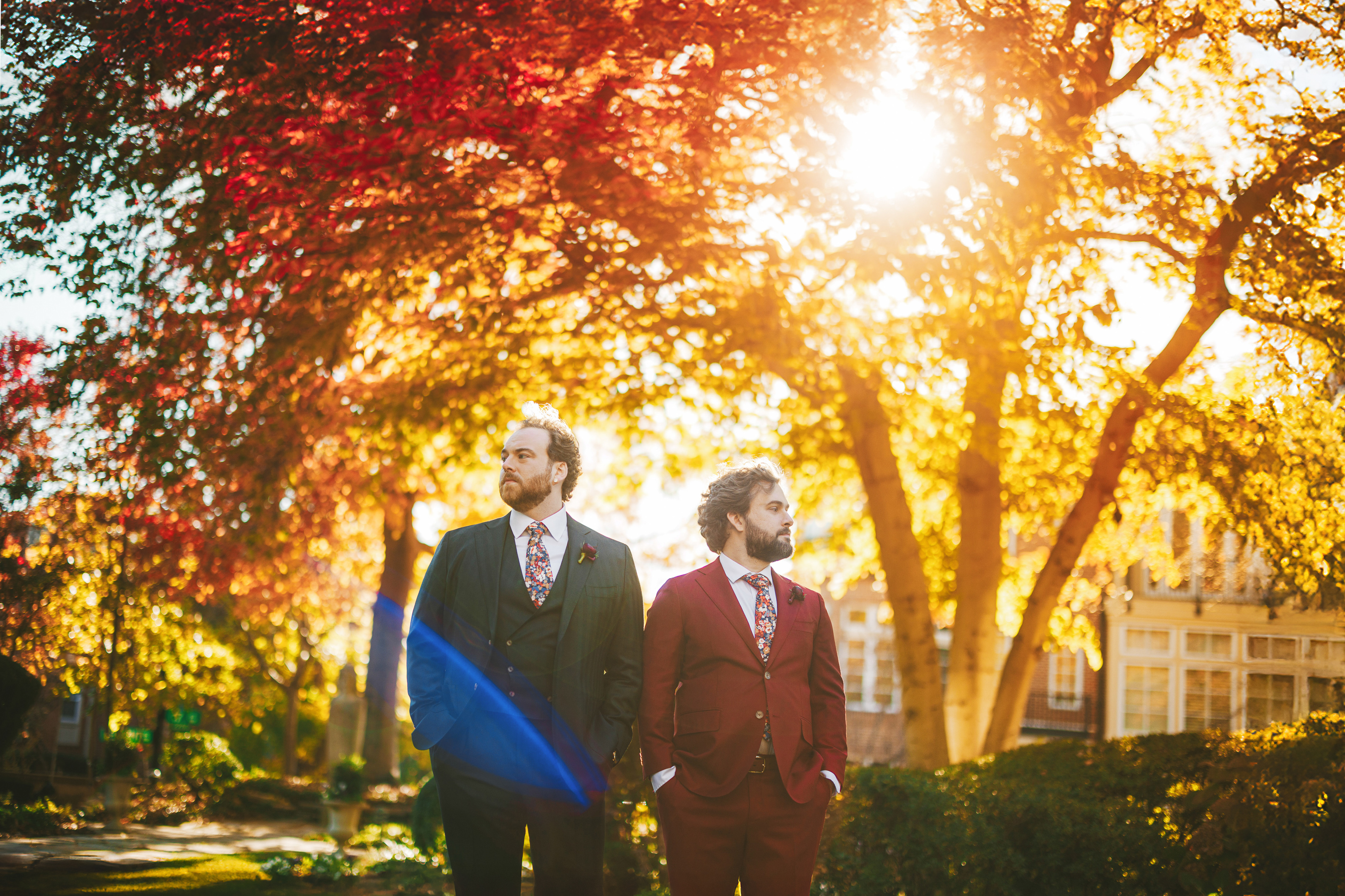 Two people in suits stand under colorful autumn trees with sunlight filtering through the leaves.