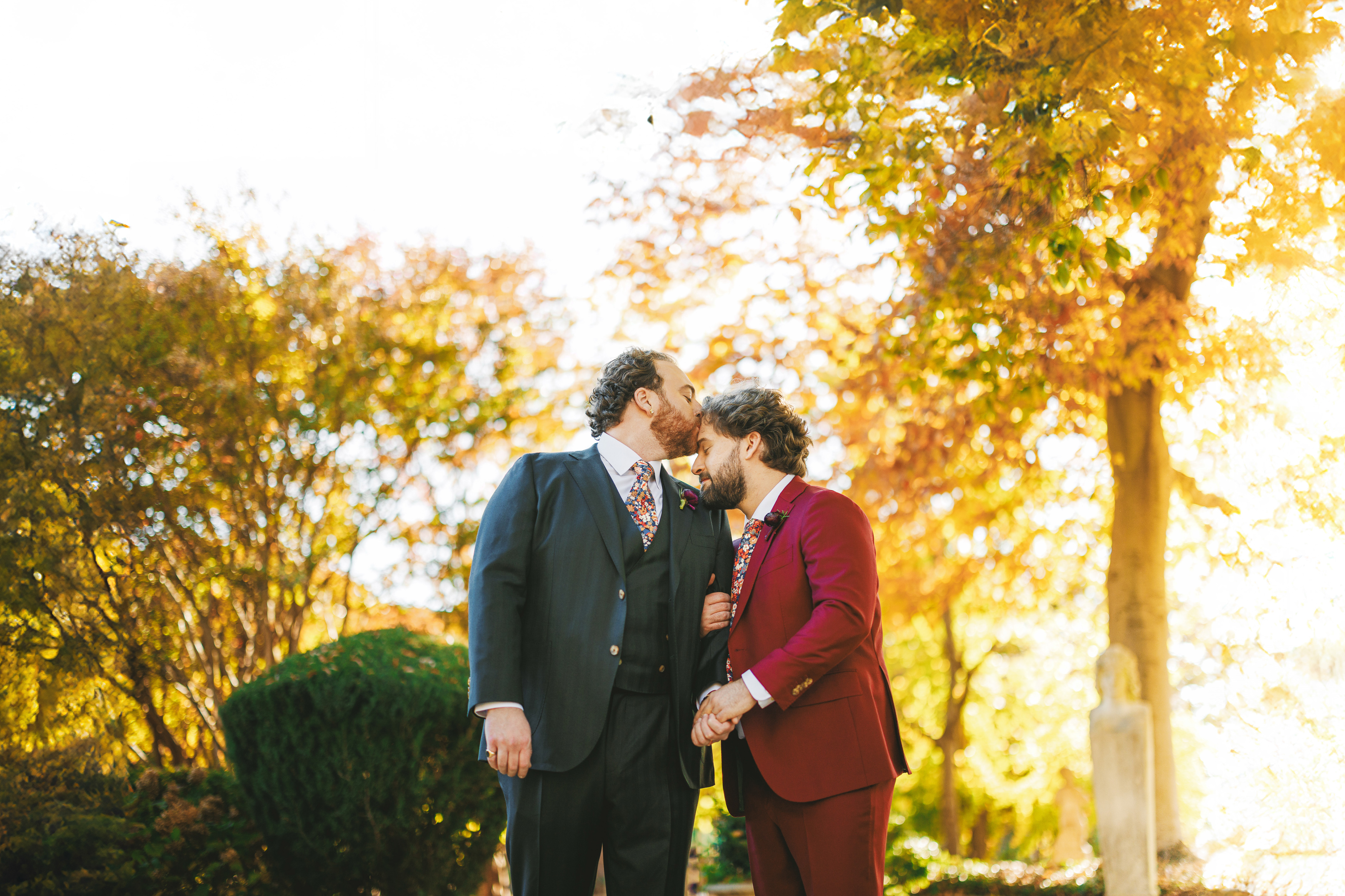 Two men in formal attire stand outdoors in a sunlit autumn setting, with one kissing the other on the forehead.