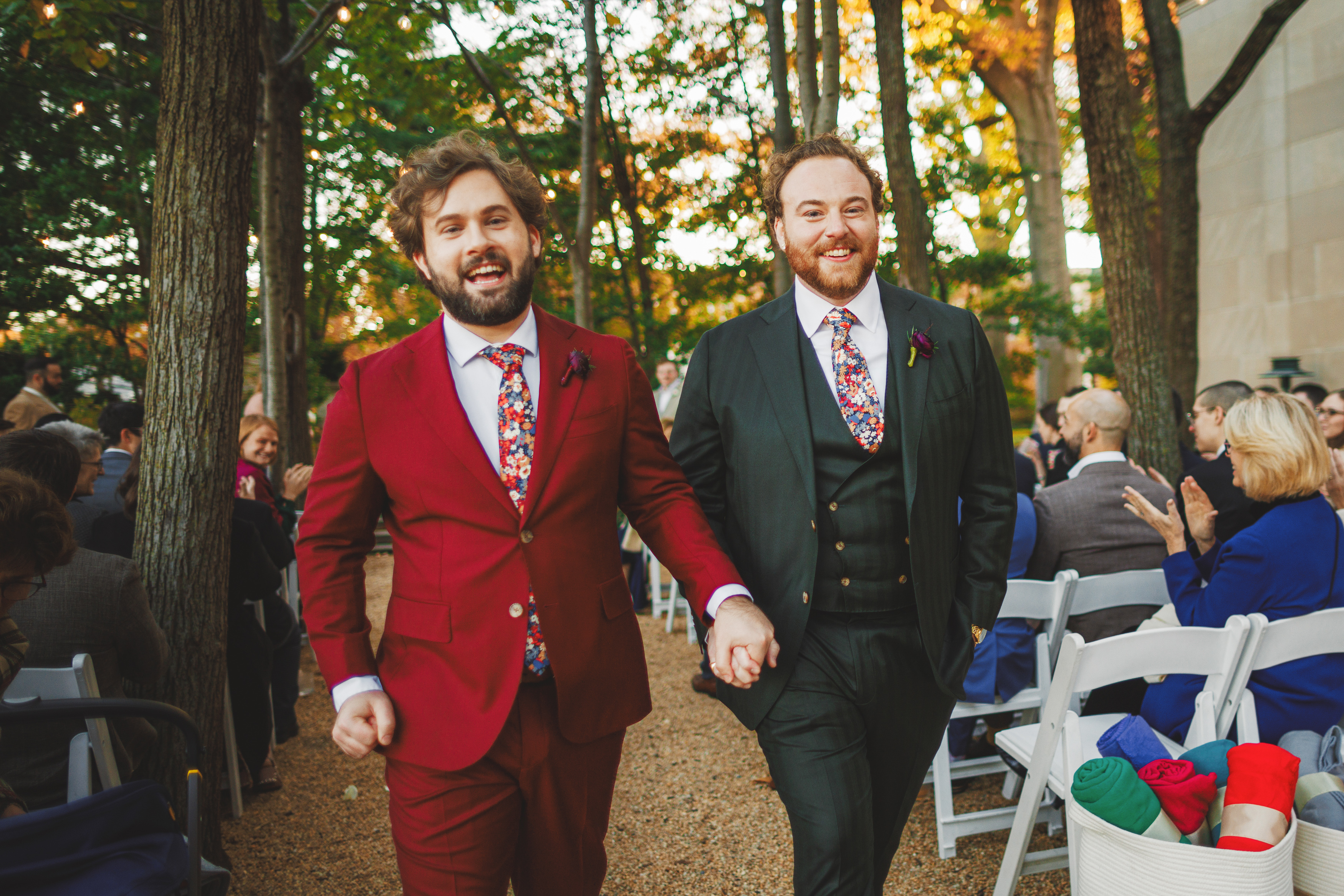 Two men in colorful suits hold hands and walk down an outdoor aisle lined with guests. One is in a red suit, the other in a dark green suit.