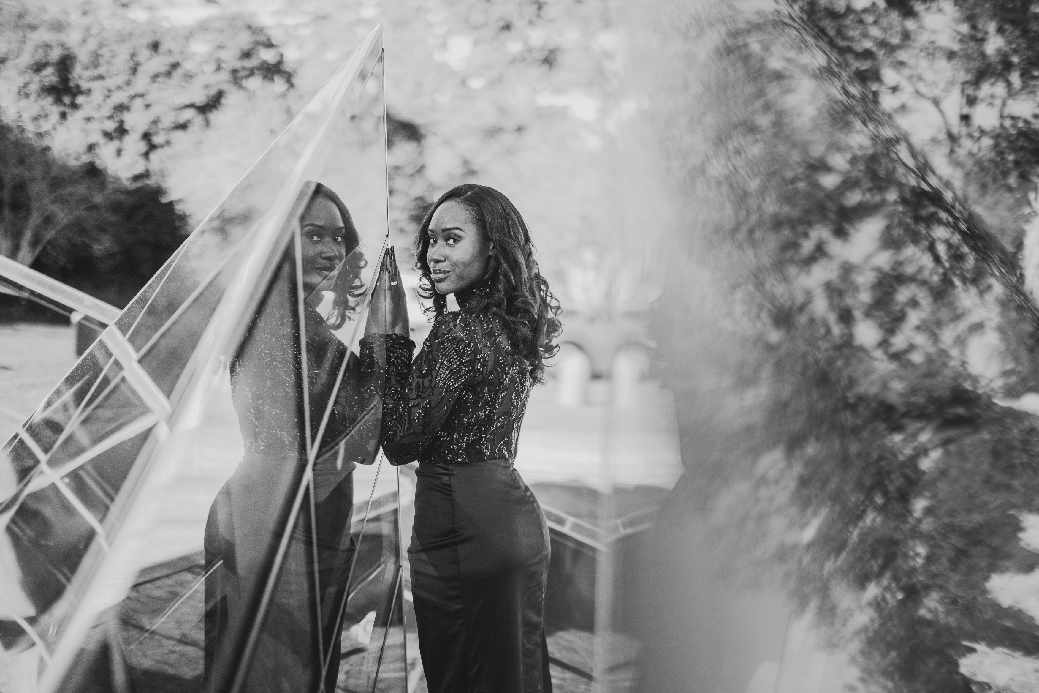 A woman in a dark outfit stands by a glass structure, with her reflection visible. The background features trees and blurred elements. The image is in black and white.
