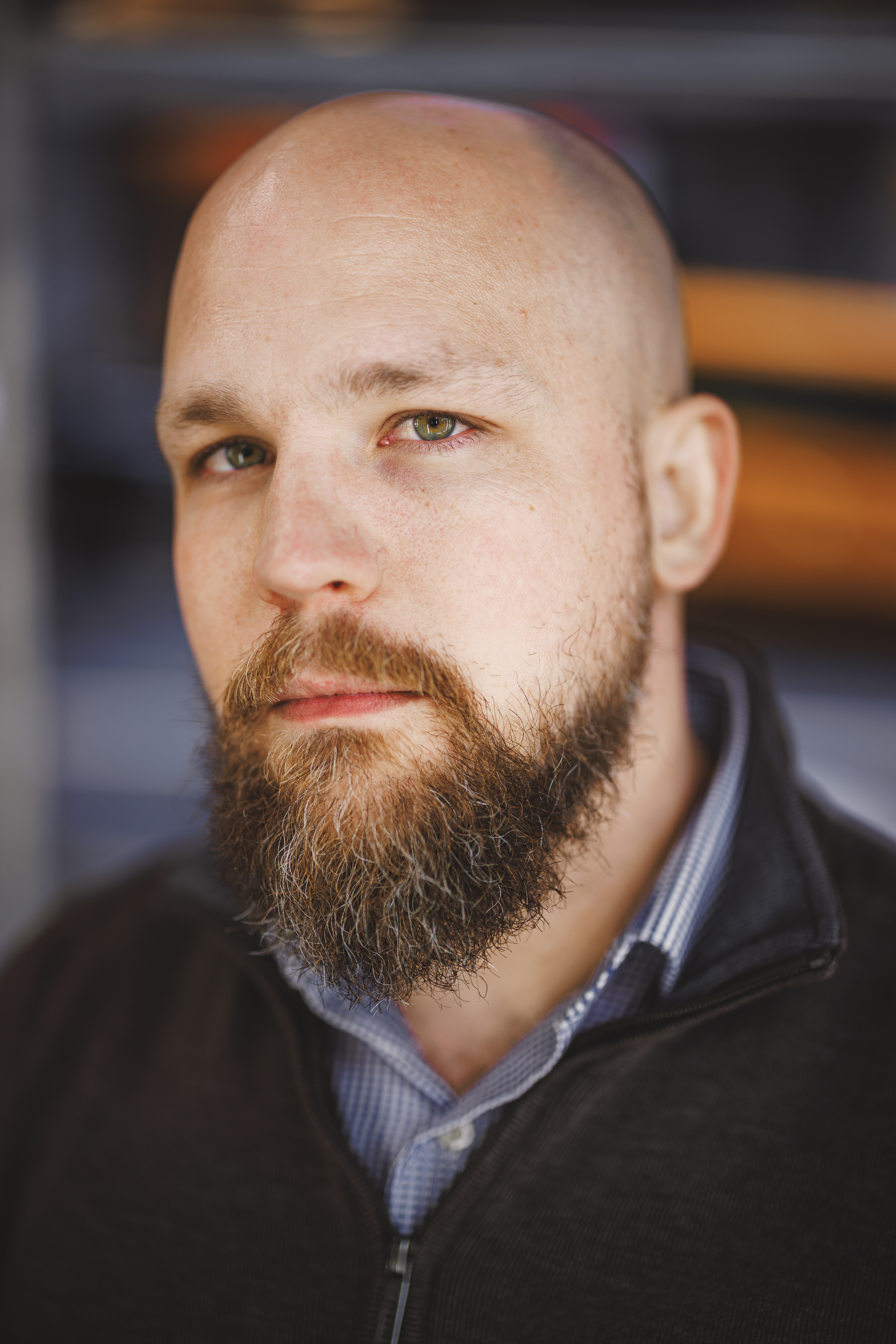 A bald man with a beard and mustache is wearing a zipped-up dark jacket over a collared shirt, looking at the camera with a neutral expression, as if ready for an AfterShoot review or some AI Editing magic.