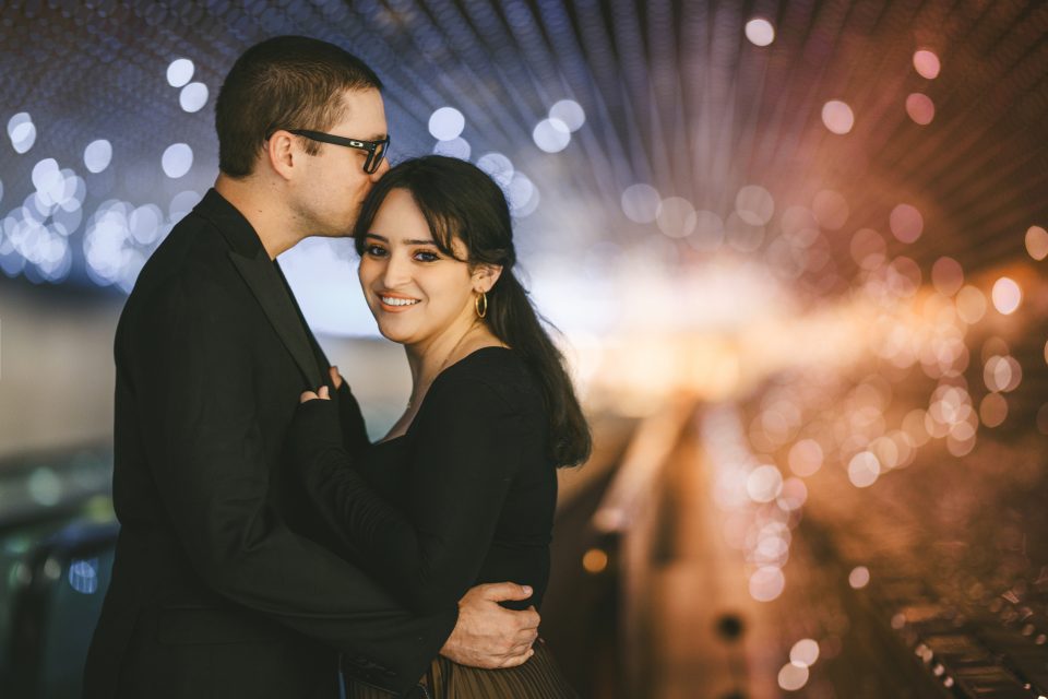 A man kisses a woman's forehead as they stand in an illuminated tunnel with bokeh lights in the background. A man kisses a woman's forehead as they stand in an illuminated tunnel with bokeh lights in the background.
