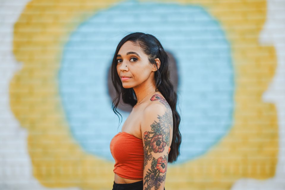 Woman with long hair and a floral tattoo on her arm stands in front of a colorful mural, wearing an orange top. Woman with long hair and a floral tattoo on her arm stands in front of a colorful mural, wearing an orange top.